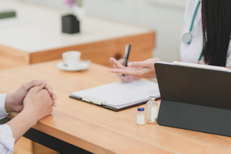 Only hands are visible in this image. A patient is opposite a doctor with a desk between them. The doctor has a laptop open and a piece of paper in front of them.